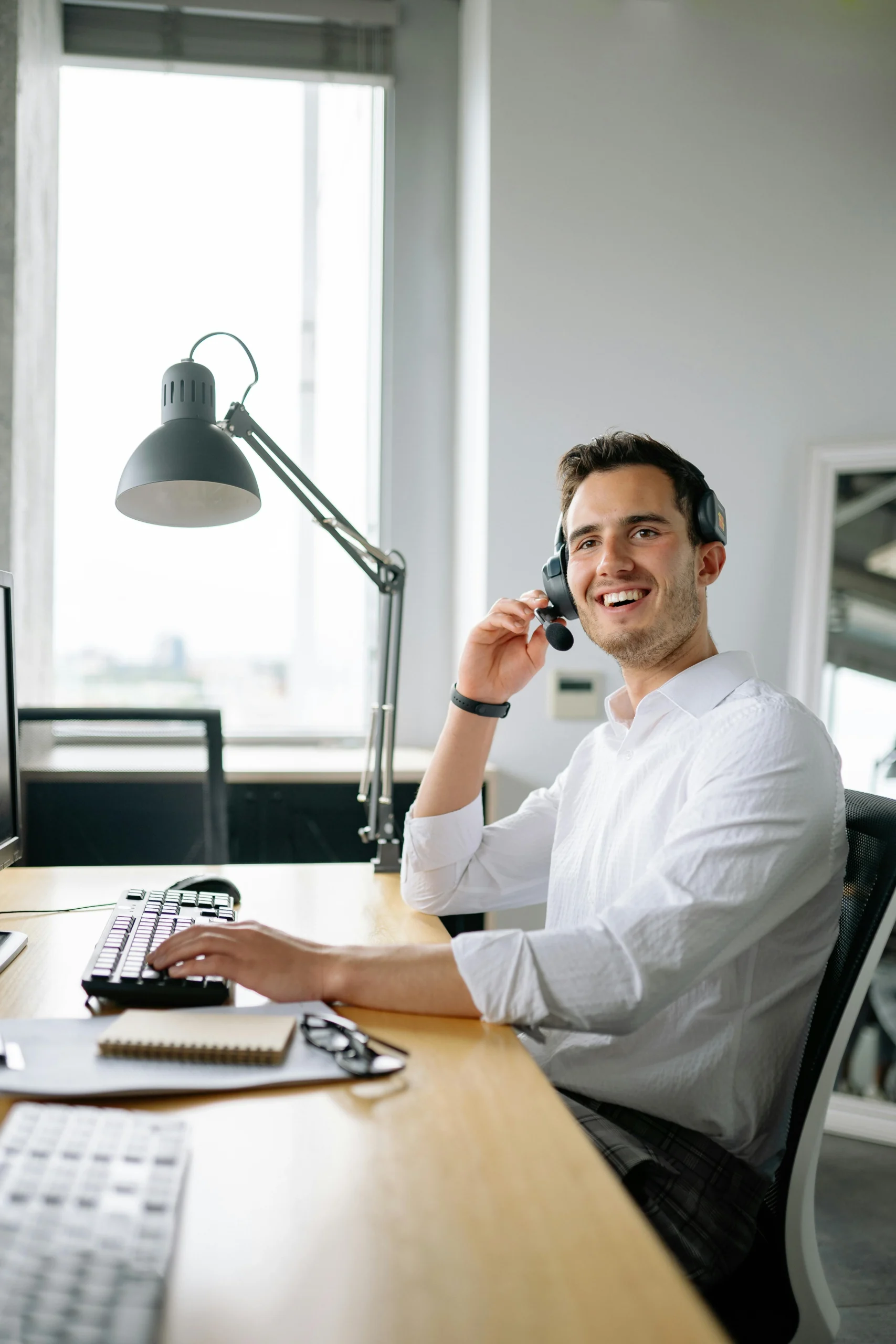Jonge man in witte blouse met headset zit aan bureau met computer, notitieboekje en bril