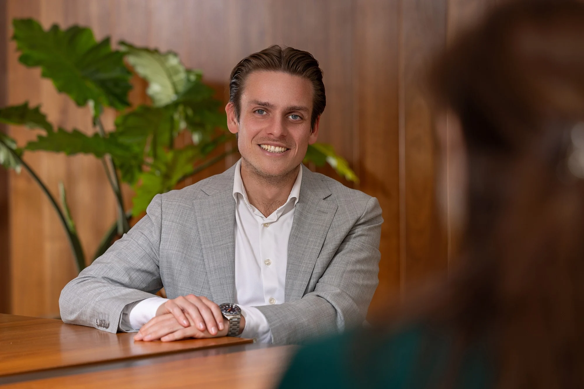Man in lichtgrijs pak met witte blouse zit aan houten tafel, groene plant op achtergrond